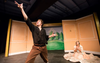 A Victorian male suitor stands reaching towards the sky professing romantic poetry in front of a female seated on the floor, looking at the suitor entranced.