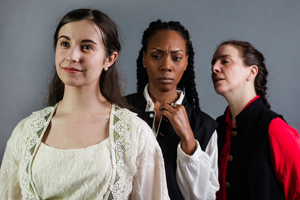 Desdemona, a woman in white smiling in front of Othello, a woman with a contemplative look on her face, in front of Iago, a woman whispering into Othello's ear.