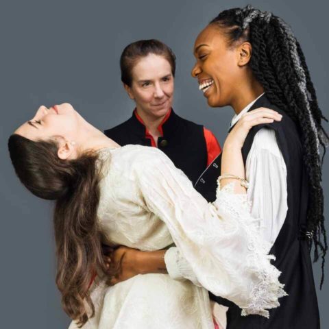A white, young woman (Desdemona) tips her head back playfully while in an embrace with her black husband (Othello) who smiles at her. Iago, smiles at the two lovers with a twinkle of envy and mischief.