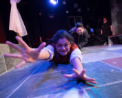 Actor laying on the floor with hands reaching out forward as another actor pulls her off stage from behind