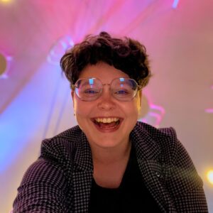 Maggie Smith, winner of the 2023-2024 Joining Sword & Pen competition. A woman with glasses and short brown hair receives the news with a large, open-mouthed smile.